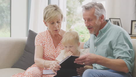 Grandparents-Sitting-On-Sofa-With-Granddaughter-At-Home-Looking-Through-Photo-Album-Together