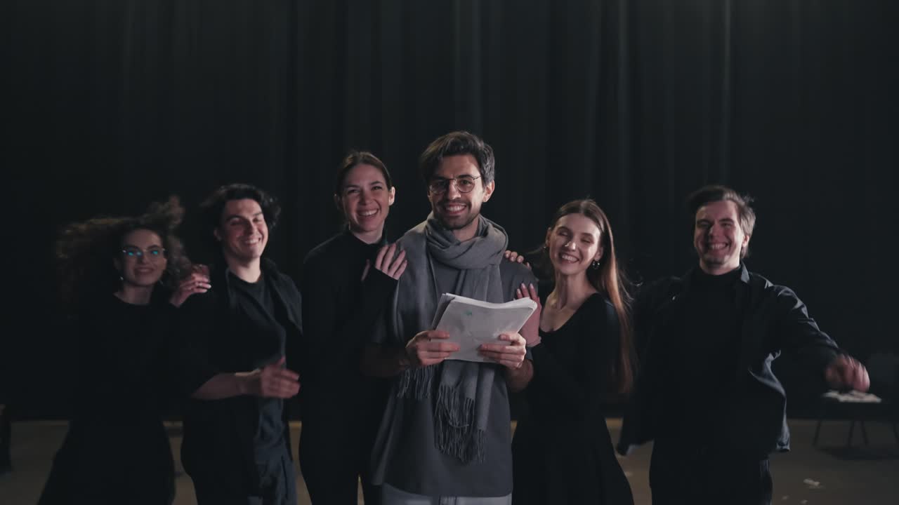retrato de un feliz joven director de teatro con una camiseta gris y una bufanda mientras los actores con trajes negros corren por detrás y abrazan a su director en el escenario de un teatro con una cortina negra