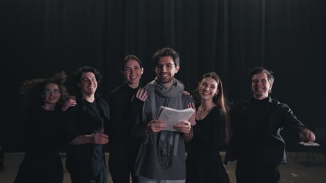 Portrait-of-a-happy-young-stage-director-in-a-gray-T-shirt-and-a-scarf-as-actors-in-black-suits-run-up-from-behind-and-hug-their-director-on-stage-in-a-theater-with-a-black-curtain