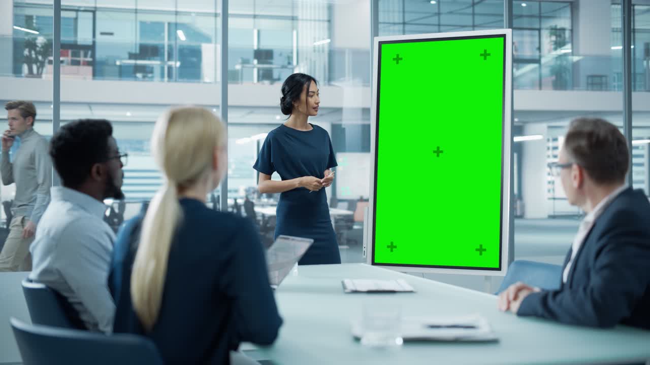 una mujer gerente de operaciones lleva a cabo una presentación de reunión para un equipo de economistas. una mujer asiática utiliza una pizarra digital con una pantalla verde vertical. la gente trabaja en una oficina de negocios.