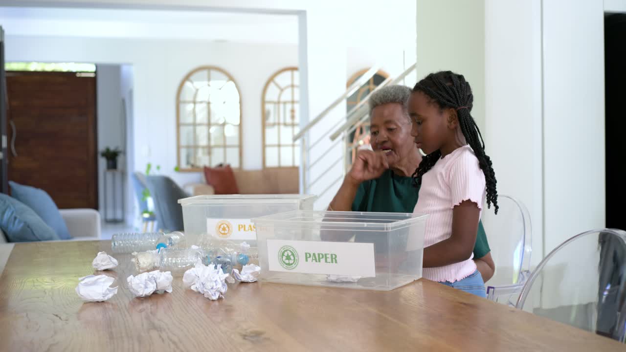 African American senior female holding paper teaching child sorting into PAPER PLASTIC bins at home