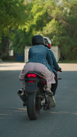 dos mujeres montan una bicicleta eléctrica en una carretera urbana. el pasajero se aferra al ciclista mientras pasan por una señal peatonal, los árboles bordean la carretera, con un poste eléctrico y un coche borroso delante de ellos