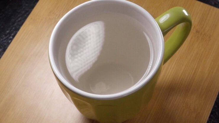 Green ceramic cup filled with clear liquid resting on wooden table with white interior and curved handle under soft indoor lighting in a kitchen setting