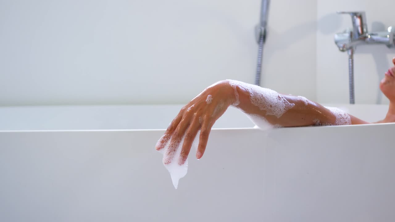 Senior African woman resting arm on white tub rim, bathing, adjusting fingers, stretching foam drip