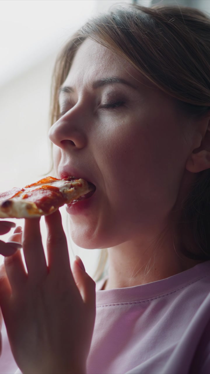 Close up of woman eating pizza. subject holds and takes bites from food with fine details like hand positioning on slice. Soft lighting creates gentle shadows in indoor setting focusing