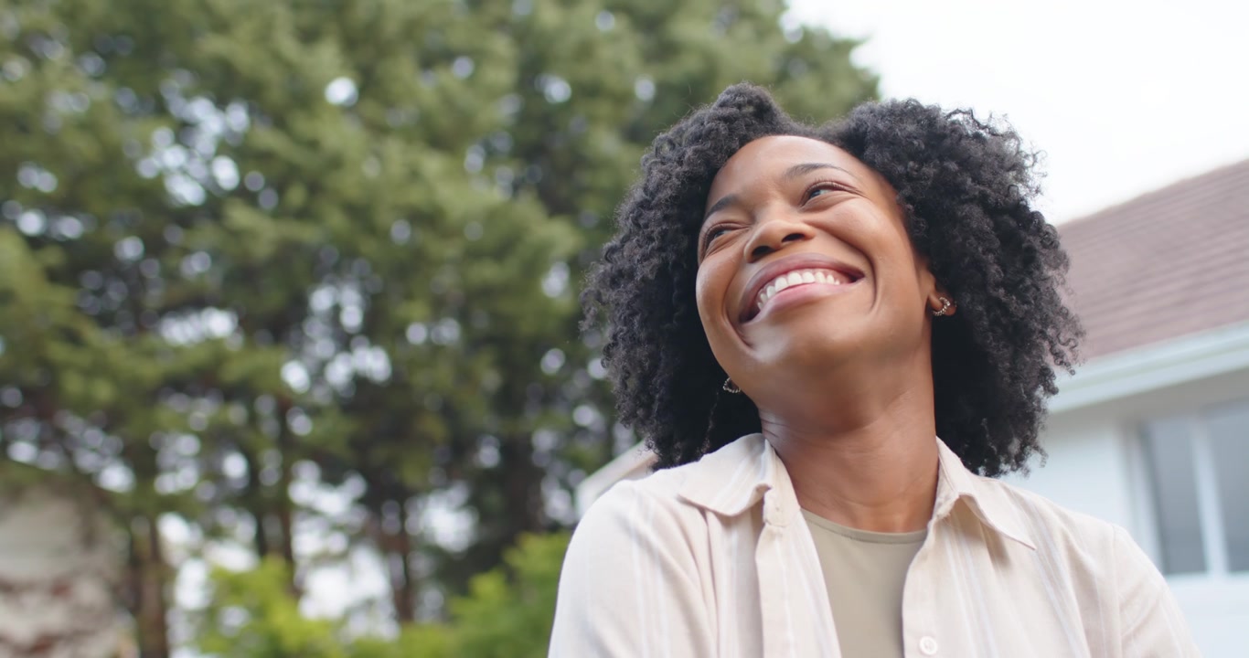 Adult African American woman looking up reacting with widening smile by house trees wearing tan top