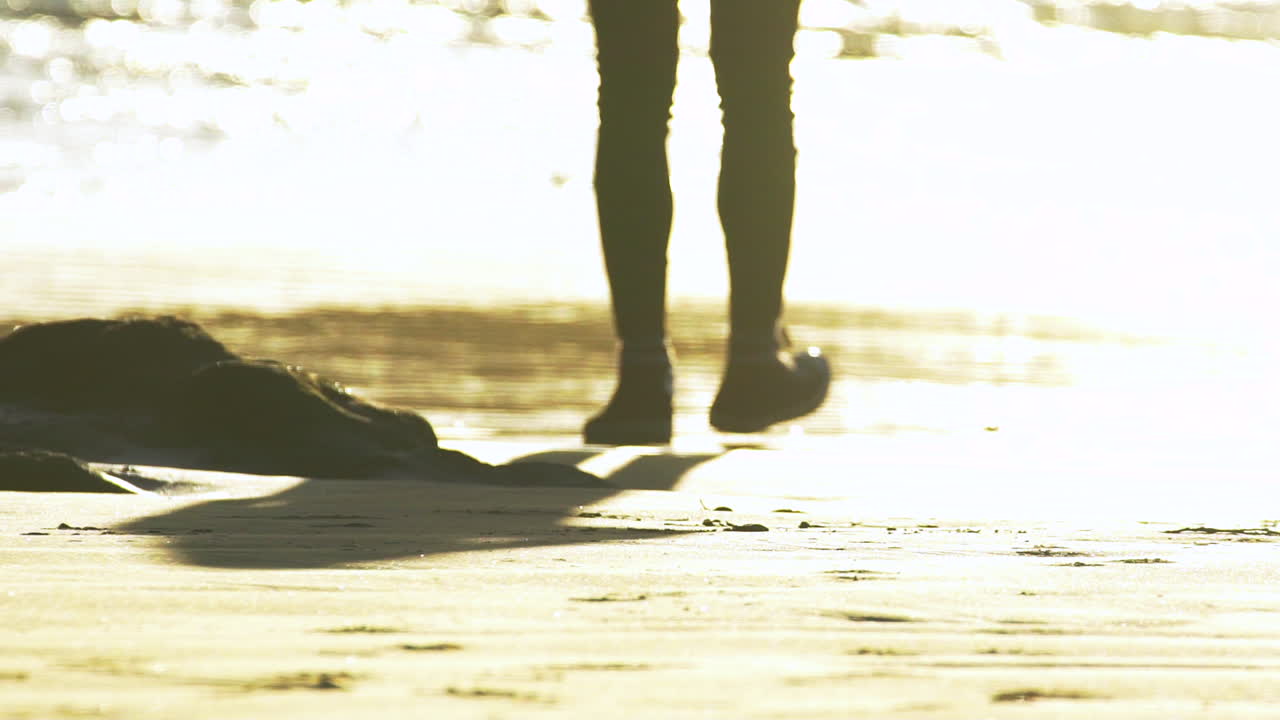 밝은 황금시간 일몰 동안 반짝이는 바닷물을 향해 느린 동작으로 걸어가는 마른 검은색 바지를 입은 여성의 다리