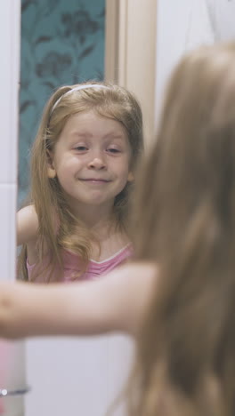 positive little blonde girl with blue eyes looks at large mirror in light modern bathroom backside close view