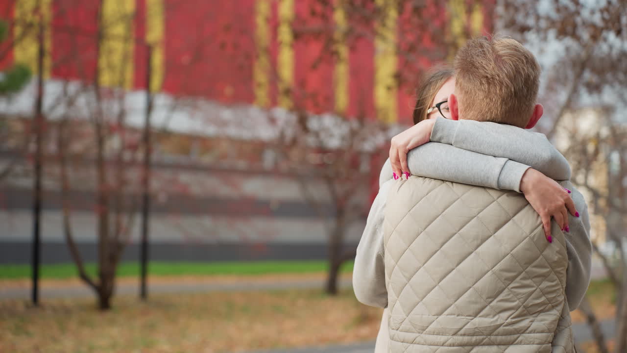 Young man smiles warmly after being hugged by sisters affectionately, standing outdoors with autumn trees swaying gently and large colorful mall in background