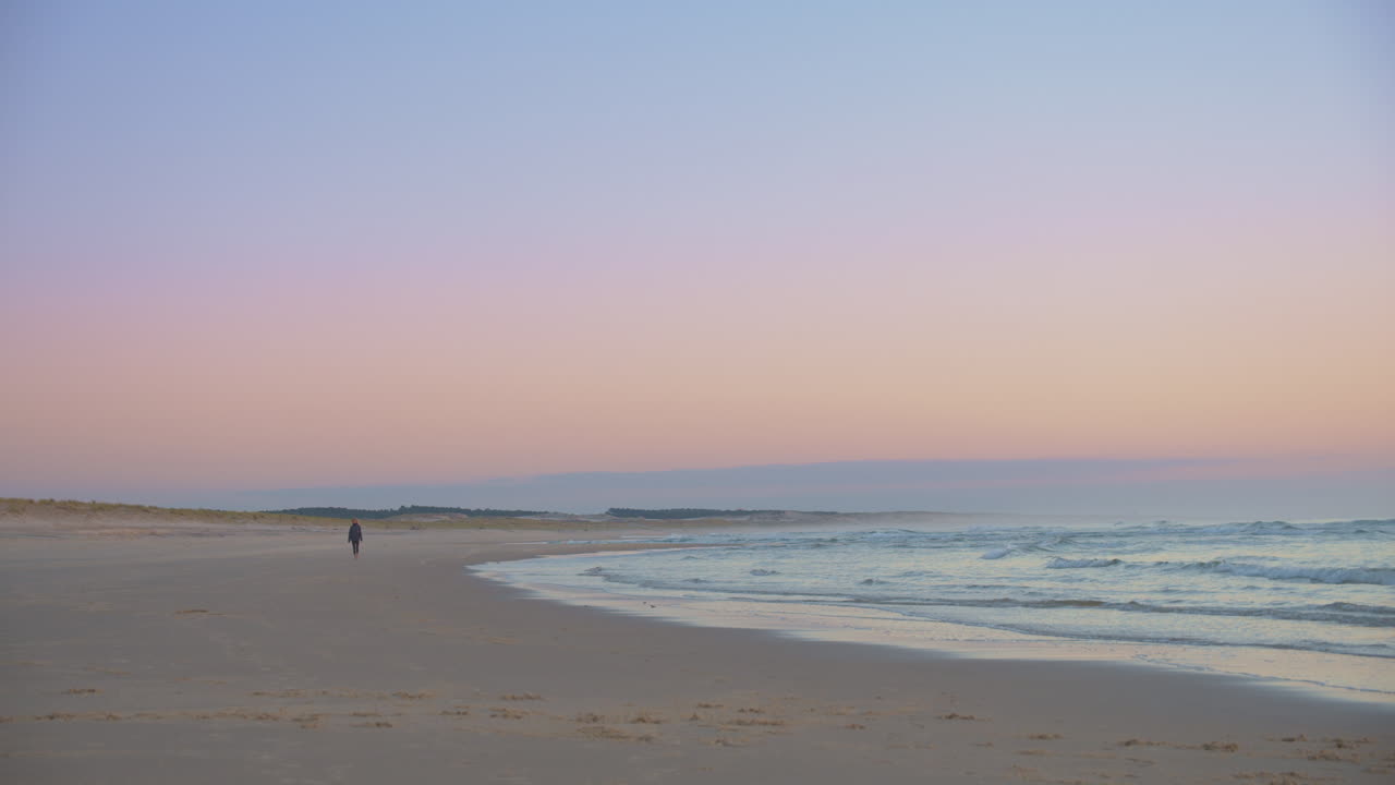 establecer una toma cinematográfica de una persona caminando sola durante una colorida puesta de sol en una playa a cámara lenta