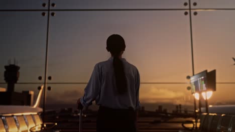 back view of asian woman with rolling suitcase waits for flight in boarding lounge at the airport, walking and looking around, airplane takes off outside the window