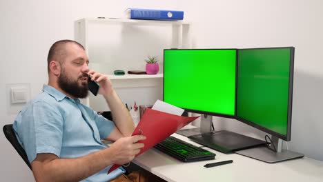 a man looks into a folder and speaks on the phone. yells at the interlocutor. a man working at home in front of two green monitors.