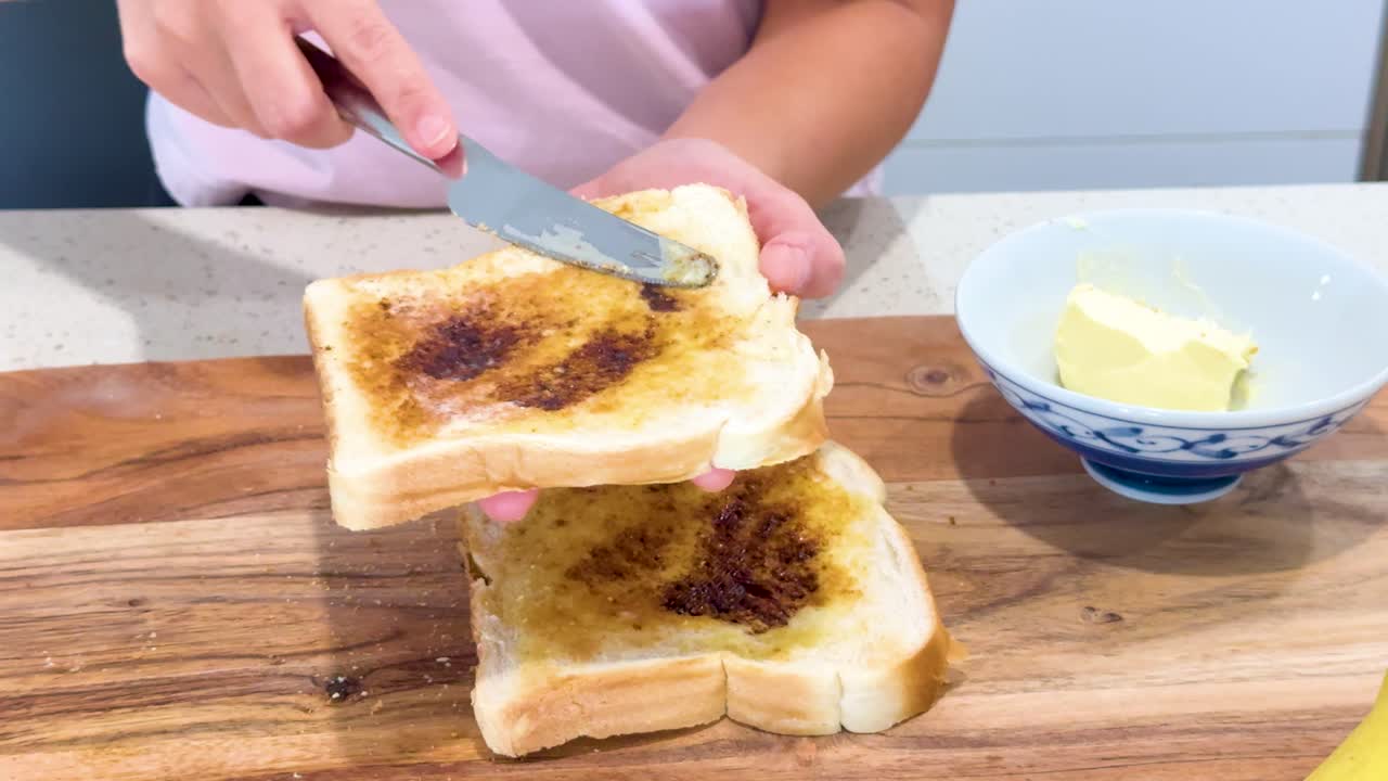 Person spreads butter on slices of toasted bread with dark yeast spread, using a knife on a wooden board in a bright kitchen setting