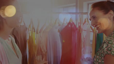 Animation-of-bokeh-lights-over-two-happy-women-talking-and-looking-at-clothes-in-shop