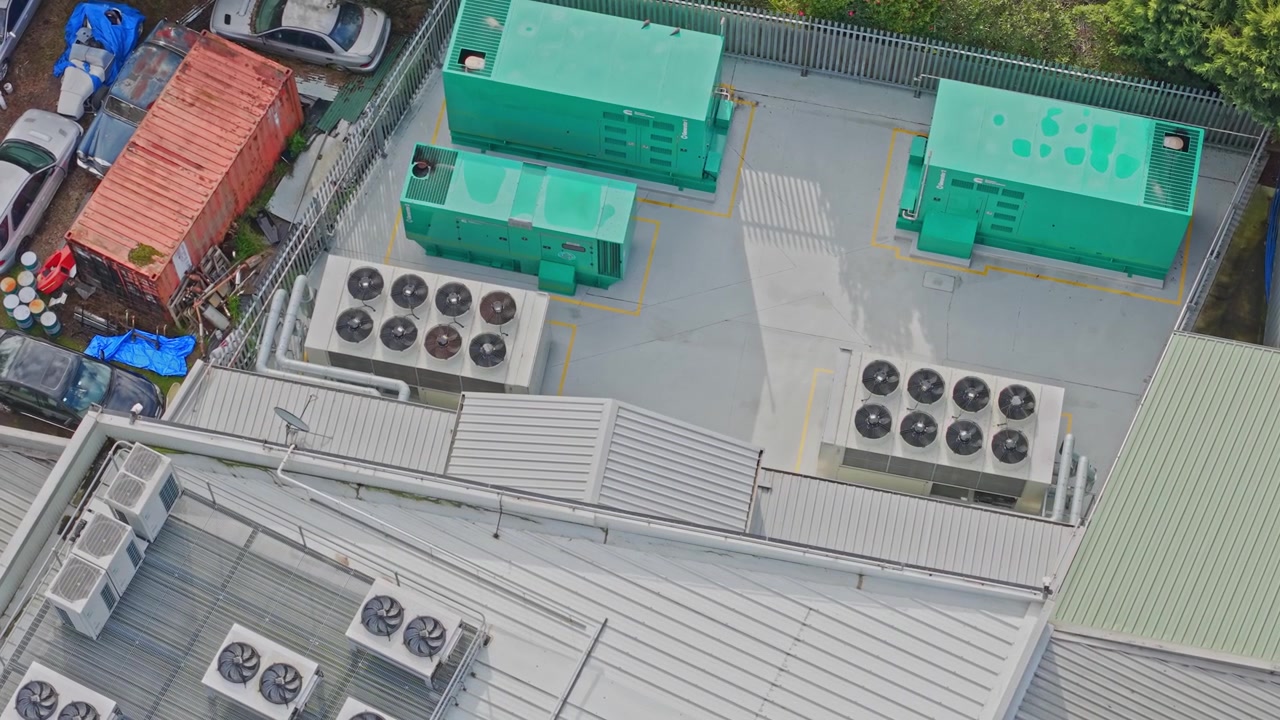 Drone shot of a data centre rooftop shows industrial generators, large HVAC units, exposed piping, and secure fencing in daylight, emphasizing modern tech infrastructure and clean composition.