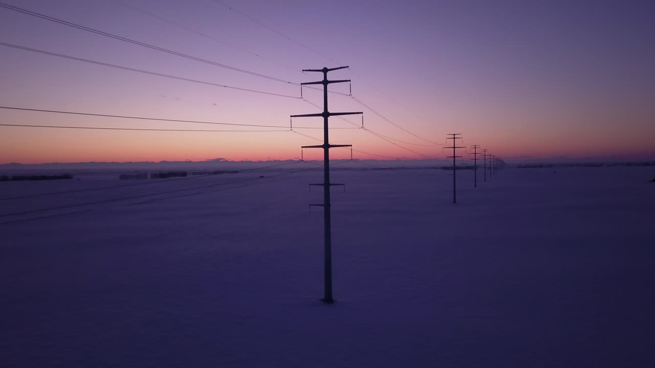 Rising tilt aerial: Modern power lines in flat snowy field at sunset