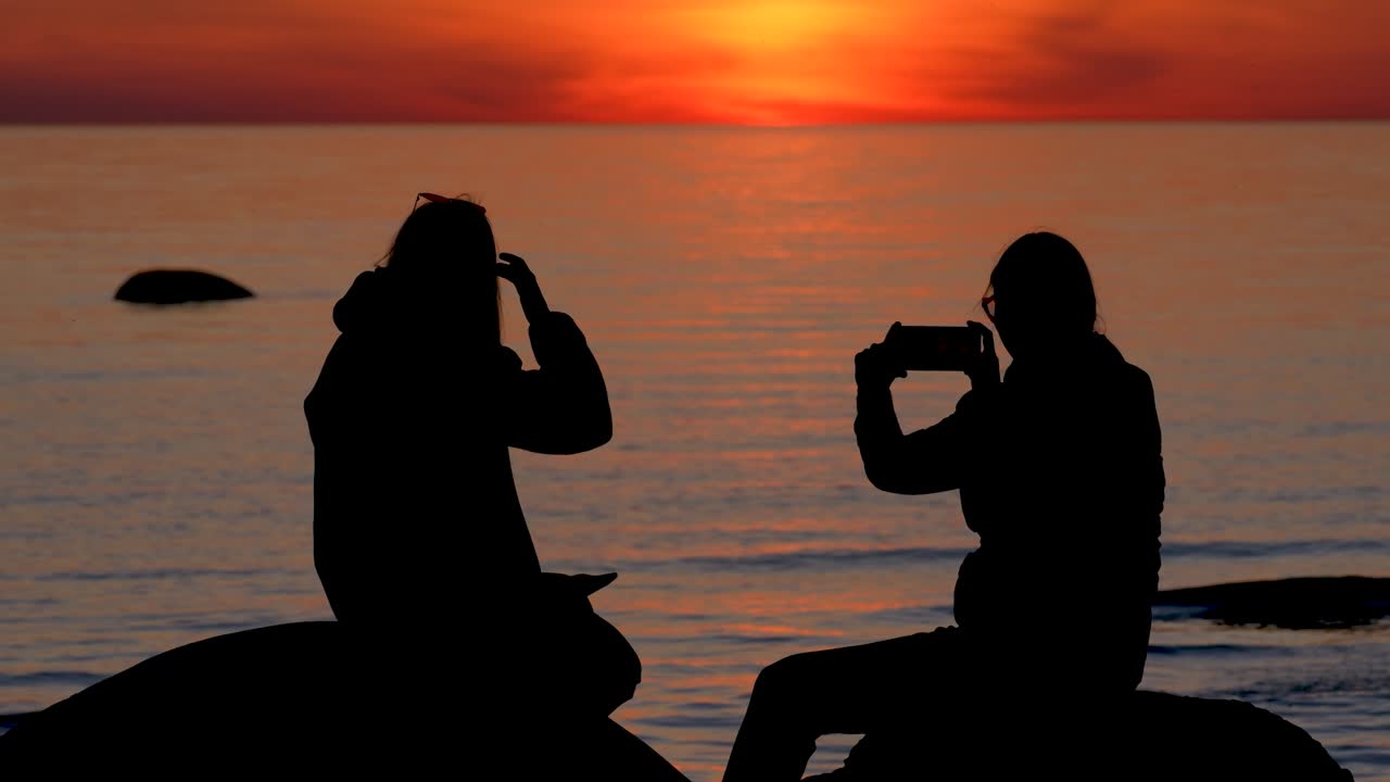 Young women silhouettes sitting by water during sunset.