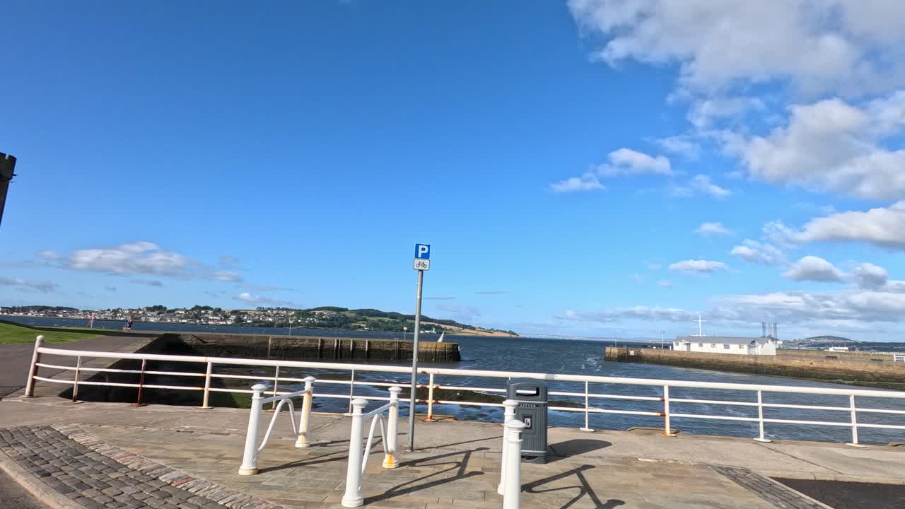 Camera moves from grassy field by stone castle toward waterfront pier, under bright daylight with clear skies, revealing coastal town and sea views