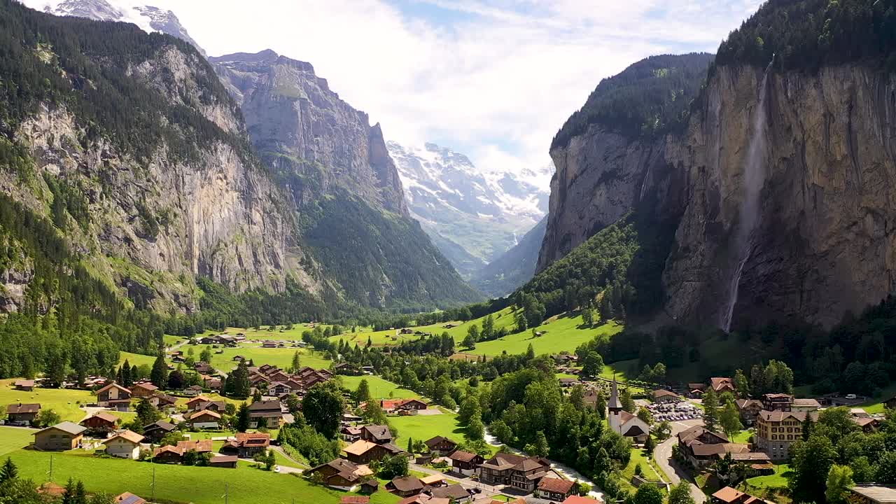 Villaggio svizzero della valle di Lauterbrunnen tra verdi prati alpini, la cascata di Staubbach e vette montuose coperte di neve.