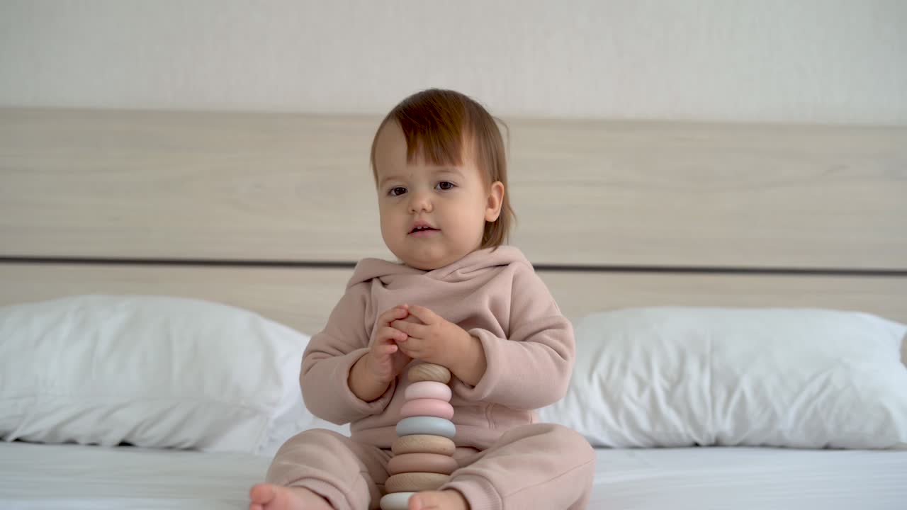 다채로운 무지개 장난감 피라미드와 함께 놀고있는 귀여운 아기 소녀가 빛 침실에서 침대에 앉아 있습니다.