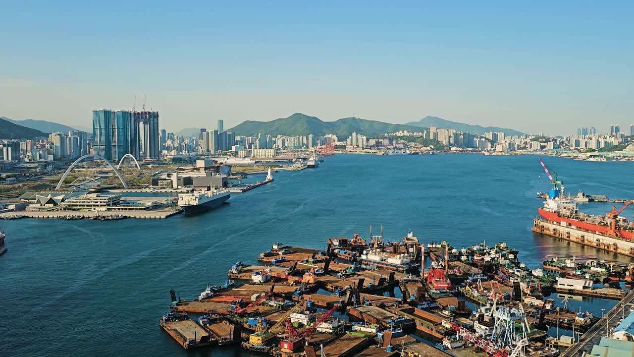 Aerial view of Busan harbor with construction barges, cranes, and modern skyline surrounded by green mountains and sea