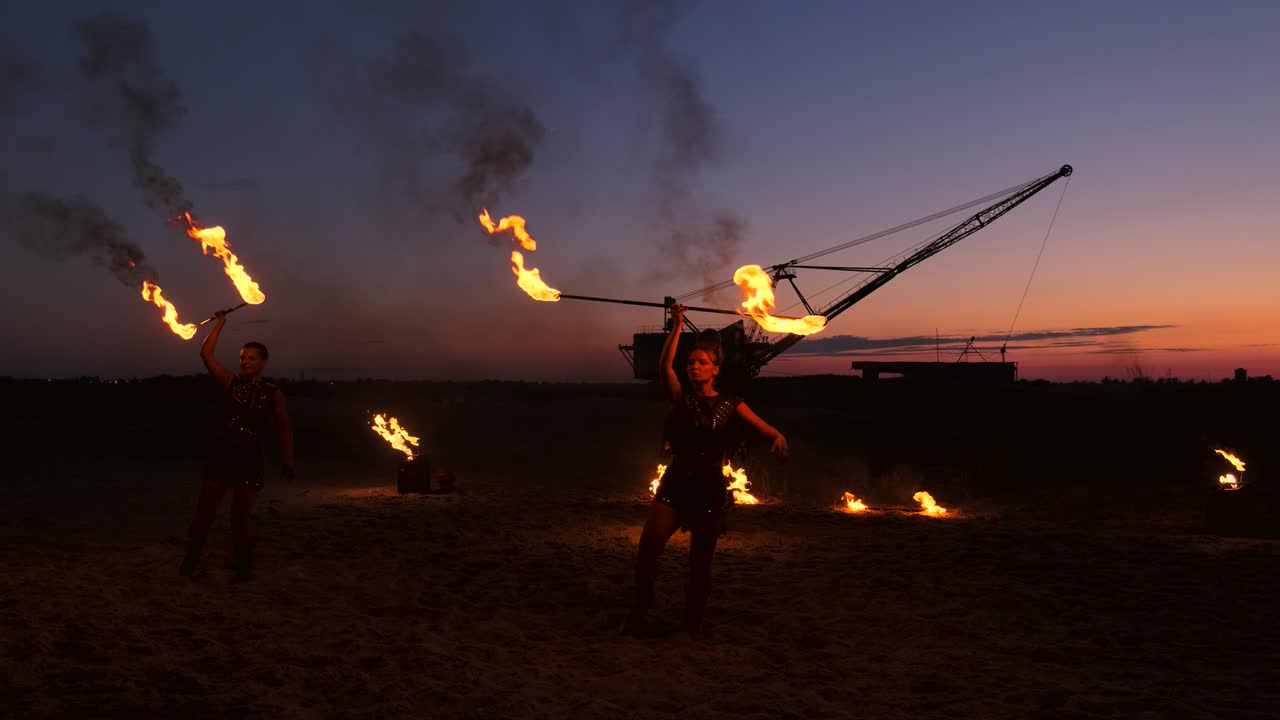 A group of men and woman fire show at night on the sand against the background of fire and tower cranes