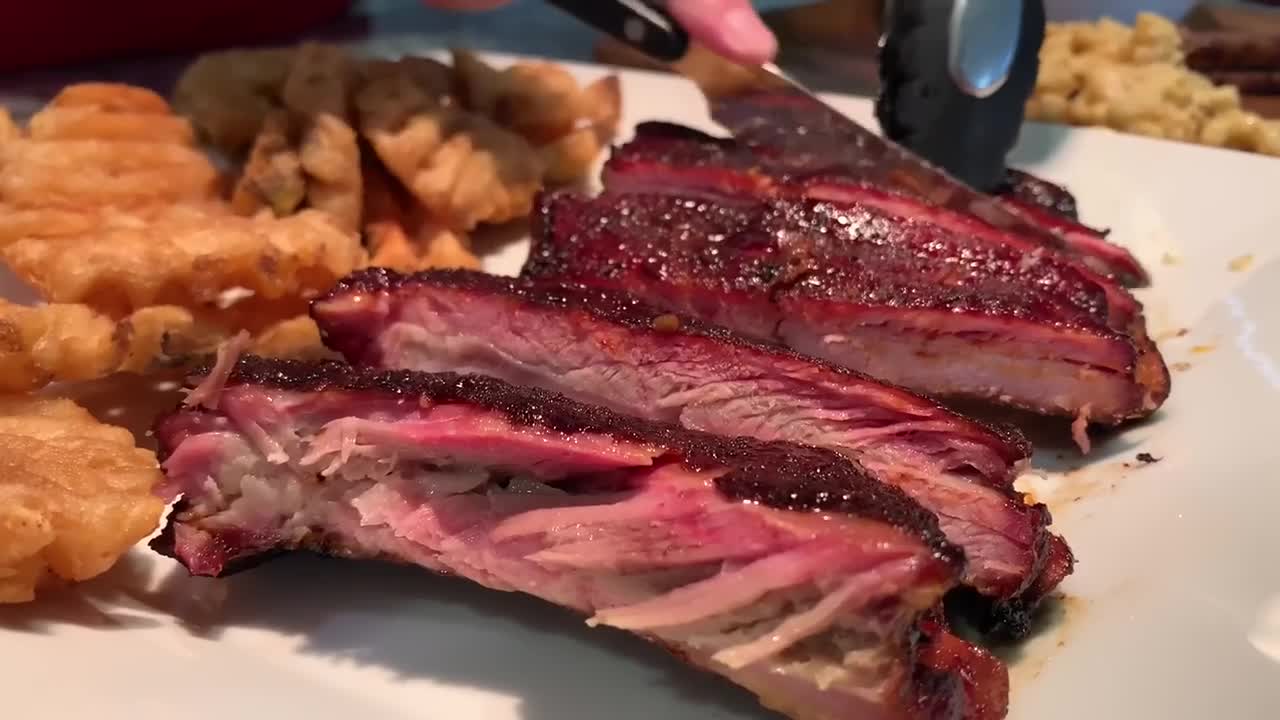 Mouth watering baby back ribs glazed over with BBQ sauce. Side of waffle cut fries. Knife cutting through the meat over a white plate. HD food stock footage video.