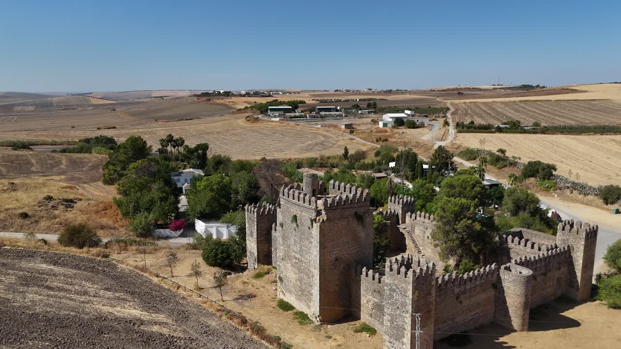 Drone in ascesa e che torna indietro sopra il Castillo de las Aguzaderas del XIV secolo, Siviglia, Spagna, che mostra mura in pietra, torri, alberi verdi e il paesaggio andaluso asciutto e soleggiato