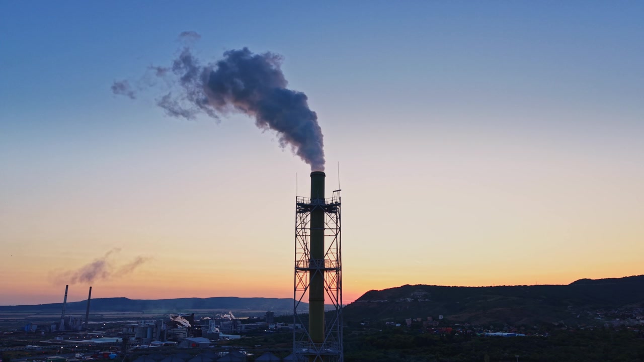 Ciminiera industriale che emette fumo durante il tramonto in un paesaggio.