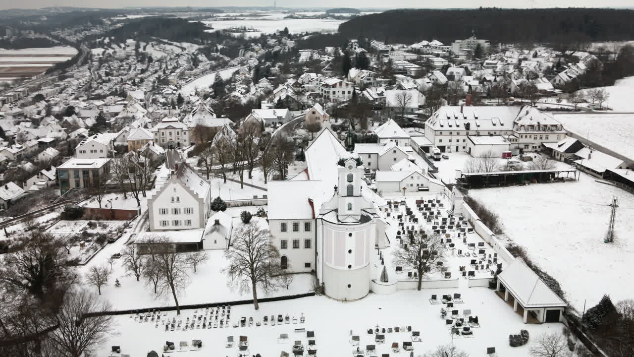 Dron obíhající zleva zasněžený kostel v Elchingenu s hřbitovem, vesnické domy, zasněžená pole pod zataženou oblohou. Ideální pro cestovní dokumenty, reklamy a odhalení