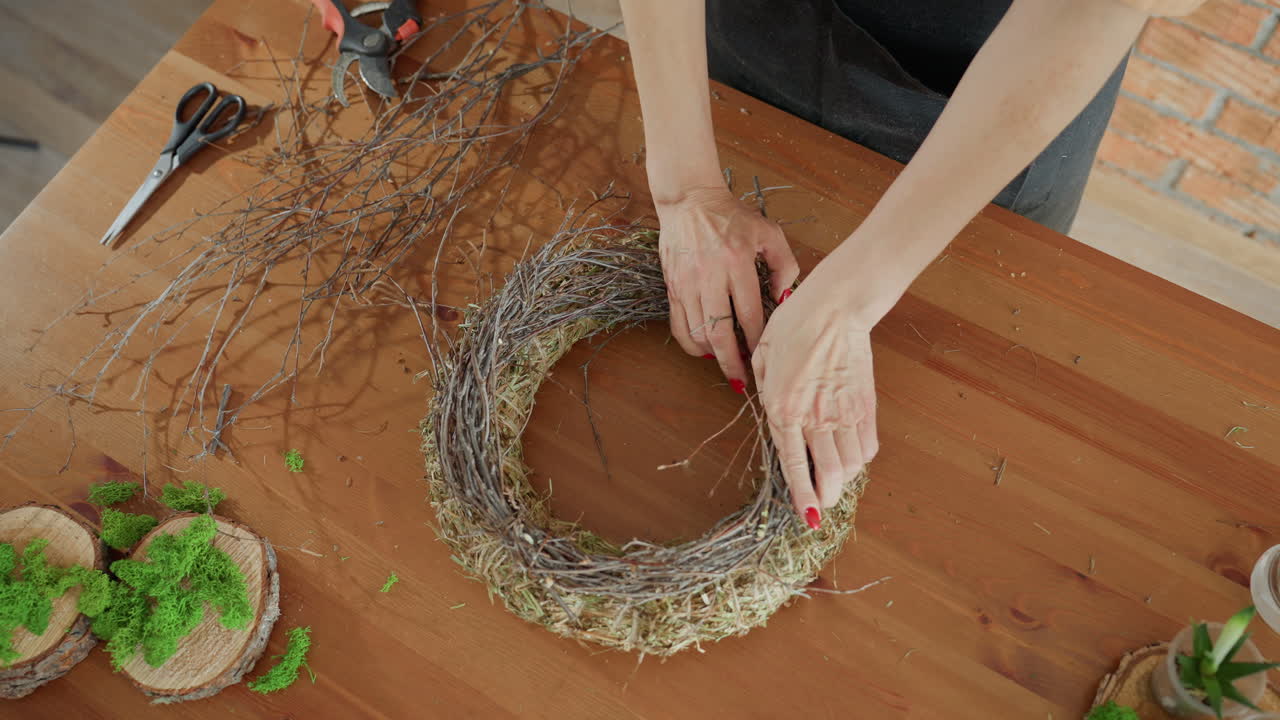 Female florist hands arranging twigs on hay wreath base on wooden table, crafting rustic handmade decoration with natural moss, wood slices, showcasing detailed artisan creative process in workshop