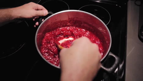 male hands stirring juicy cranberries in saucepan boiling sauce on kitchen stove