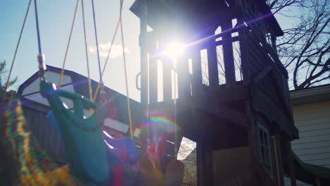 dolly past a swingset and old wooden play structure as an anamorphic sun flare peeks through the gaps of the structure