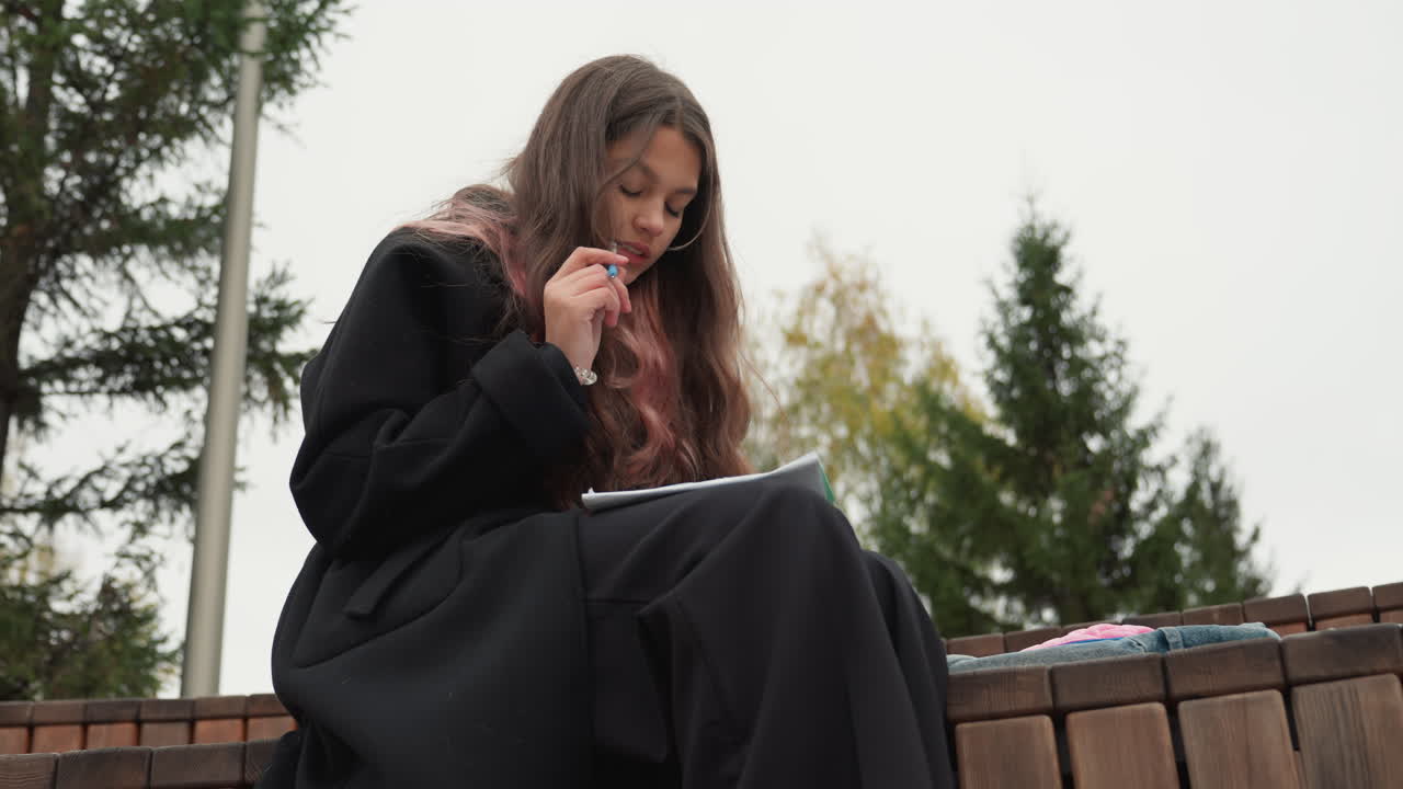 Student som skriver i notatbok utendørs, sittende på en benk under en overskyet himmel, iført en mørk jakke og med langt krøllete hår, fokusert på notater med penner og lærebøker i nærheten, reflekterende høststemning på campus, stille studie