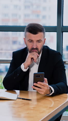 thoughtful businessman focused on the screen of his smartphone. busy man scrolling information in his phone sitting at the desk. blurred backdrop. vertical video