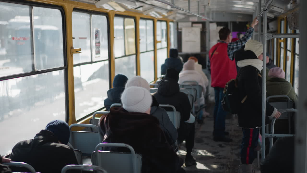 high angle interior view of crowded train during winter commute, passengers seated while others stand gripping pole as carriage slows toward stop, cold light through windows and metal frames