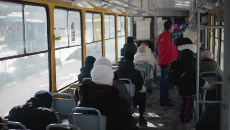 high angle interior view of crowded train during winter commute, passengers seated while others stand gripping pole as carriage slows toward stop, cold light through windows and metal frames