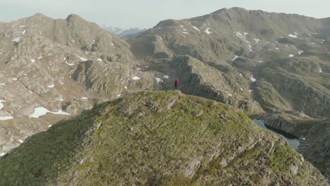 Drone-shot-of-a-man-standing-atop-a-scenic-mountain-hill-overlooking-a-vivid-blue-alpine-lake-in-the-Alps-below