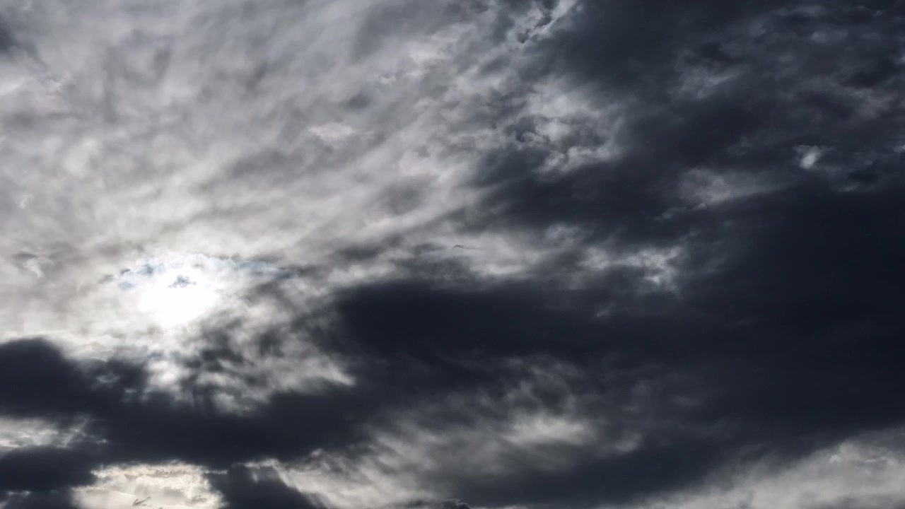 Un timelapse meteorológico de nubes de tormenta oscuras y cambiantes que destacan patrones climáticos activos. El sol permanece oculto, pero rayos de luz brillantes atraviesan el cielo pesado y nublado.