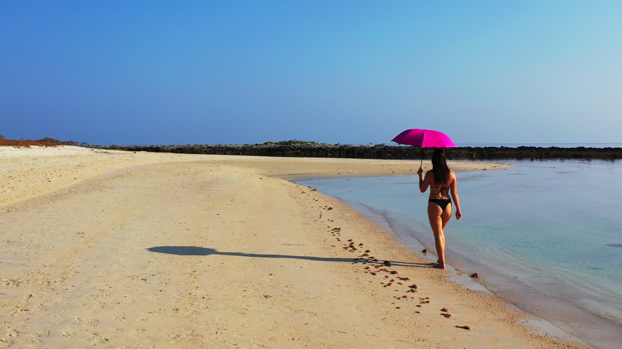 Attractive woman in swimsuit with pink umbrella walking on paradise exotic beach washed by calm clear sea water on bright blue sky background in Indonesia