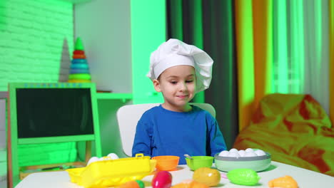 lovely toddler boy wearing white chef's cap sits at the desk. cute child cheerfully plays with grocery and plastic products.