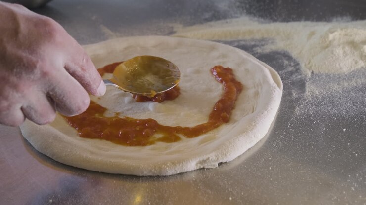 Primo piano di uno chef che spalma la salsa sull'impasto della pizza su un bancone da cucina di un ristorante.