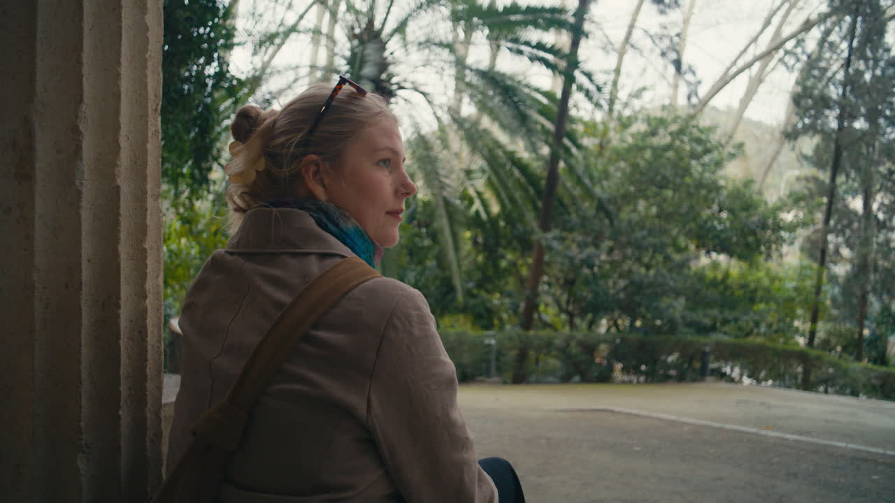 Young woman sitting near a column in a botanical garden, looking out over palm trees and greenery. Calm, reflective mood in a natural setting