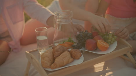 close-up shot of hand reaching for fresh green grapes from a picnic spread featuring croissants, oranges, peaches, grapes, and water with herbs in glass pitcher