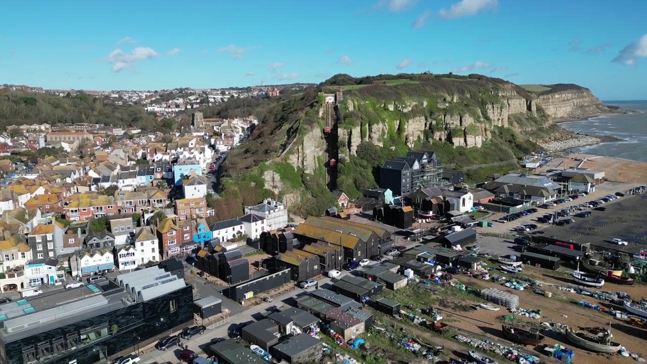 Ripresa aerea con drone di Hastings, Regno Unito, telecamera che sorvola la spiaggia di Stade, barche da pesca e la via principale, verso la funicolare di East Hill. Ripresa in allontanamento