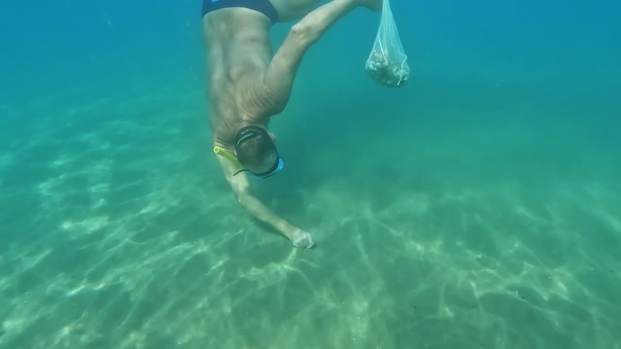 vista submarina dividida del pescador buceando con máscara y snorkel en busca de almejas navajas escondidas en el lecho marino mientras sostiene una bolsa llena de mariscos en agua de mar cristalina