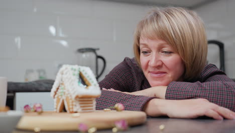 woman leans on table, thoughtfully admiring her handmade gingerbread house, cozy kitchen setting with warm lighting, potted plant, and homemade treats