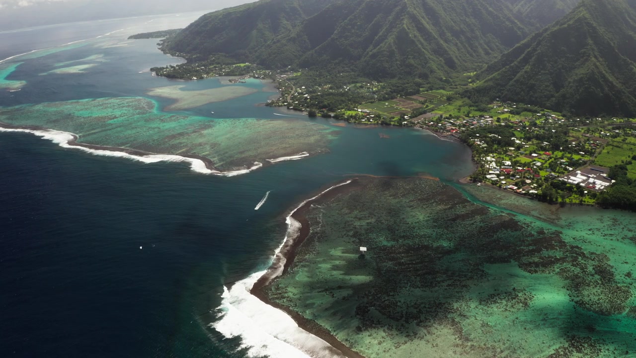 Wide aerial drone shot over Teahupoo village, Tahiti, coastal houses lining the shore as a wave breaks along the coral reef just offshore.