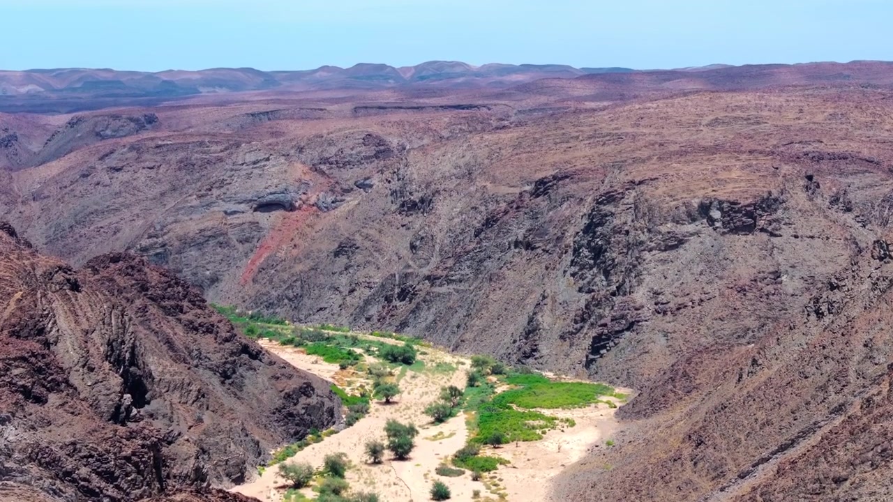 Drone-lento kasvillisuuden peittämän joenuoman yllä, joka mutkittelee karujen Namibian vuorten läpi, pieni asutus sijaitsee vihreän laakson pohjalla.