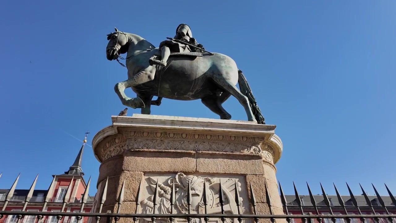Riprese a mano panoramicano l'iconica statua equestre di Filippo III nella Plaza Mayor di Madrid sotto cieli azzurri e luminosi, mostrando angolazioni dinamiche, architettura storica e la nitidezza della luce diurna.